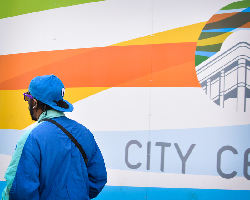 A Black man with a backward ballcap and sunglasses walks away from us, past a wall with colored stripes and partial lettering "City C..."