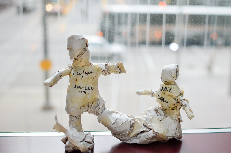 Two paper figures in front of a blurred street, one taller with chest bearing the text "I AM/NOT SWOLLEN WITH" one smaller with text "BABY BIRDS."